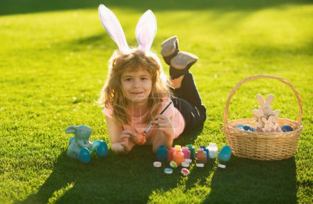 Téléchargez les photos : Enfant avec des œufs de Pâques et des oreilles de lapin pondant sur de l'herbe peignant des œufs. Joyeux Pâques enfants visage - en image libre de droit