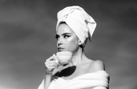 Foto de Atractiva mujer sexy sosteniendo una taza con café y mirando al amanecer. Mujer de belleza con toalla en la cabeza oliendo café en la noche en el cielo dramático al aire libre. Spa matutino - Imagen libre de derechos