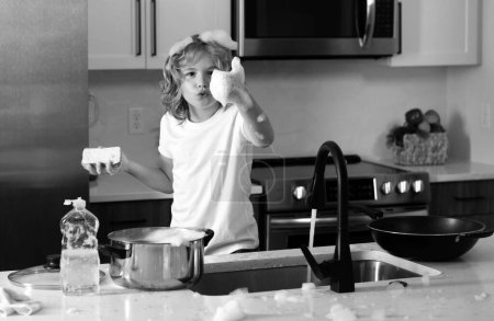 Téléchargez les photos : Petit garçon qui lave la vaisselle dans la cuisine intérieure. Nettoyer la vaisselle, le liquide vaisselle avec de la mousse. Enfant aidant ses parents avec les travaux ménagers - en image libre de droit