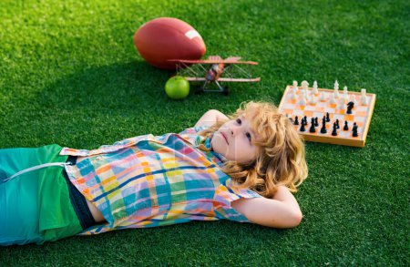 Téléchargez les photos : Enfants de rêve et concept d'enfance. Détendez-vous dans le parc, allongé sur l'herbe, rêvant. Un gamin qui pense aux échecs sur l'herbe dans un parc d'été. Le concept d'apprentissage et d "éducation des enfants - en image libre de droit
