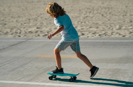Téléchargez les photos : Kid skateboarder monter un skateboard dans la rue. Enfant dans une ville d'été. Petit enfant, tout-petit garçon à cheval skateboard dans le parc pour la première fois, essayer le skateboard - en image libre de droit