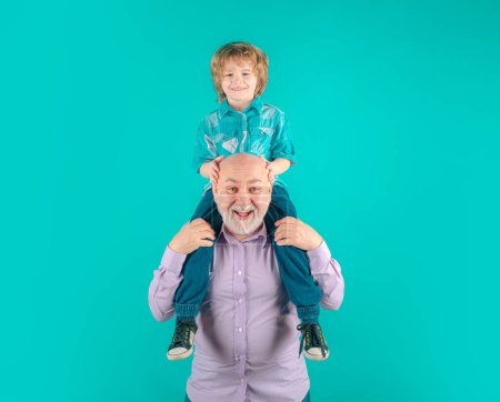 Téléchargez les photos : Jeune petit-fils et vieux grand-père piggyback avec drôle de visage isolé sur bleu en studio. Grand-père et mignon garçon avec un visage drôle - en image libre de droit