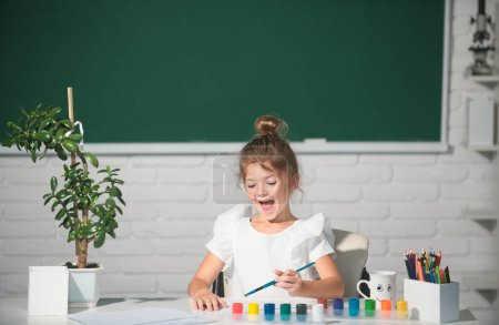 Foto de Niña dibuja en el aula sentada en una mesa, divirtiéndose en el fondo de la pizarra escolar. Educación creativa para niños - Imagen libre de derechos