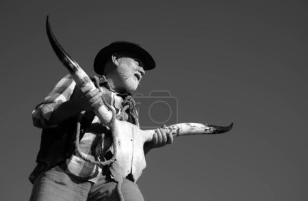 Foto de Viejo vaquero salvaje del oeste sosteniendo cráneo de vaca. Hombre occidental mayor con barba y chaqueta marrón, sombrero. esqueleto de hueso de búfalo - Imagen libre de derechos