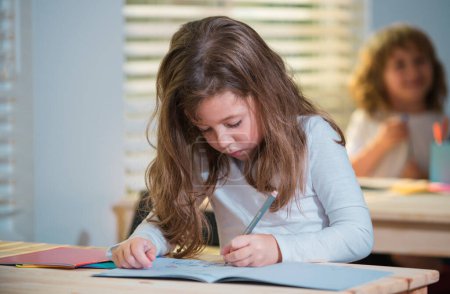Photo for Education, learning and children concept. Little student girl pupil with book writing school test. Back to school. Happy cute child is sitting at a desk indoors. Child is learning in class - Royalty Free Image