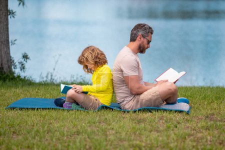 Téléchargez les photos : Père et fils aiment passer du temps de qualité à lire des livres à l'extérieur. Papa et enfant fils profiter de la nature tout en lisant le livre à l'extérieur. Père et son petit fils lisent un livre dans le parc. Fête des pères - en image libre de droit