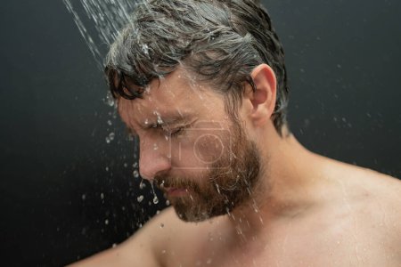Téléchargez les photos : Homme lavant les cheveux dans la salle de bain. Pommeau de douche dans la baignoire. Visage masculin sous la douche. Un homme prend une douche dans la salle de bain. Un mec qui se douche. Concept de salle de bain. L'homme est sous l'eau gouttes dans la salle de bain - en image libre de droit