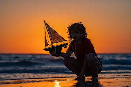 Photo for Silhouette of kid playing with toy seailing boat on sunset sea. Dream of summer. Kid play on the beach on a sunny day. Little sailor play with sailing boat in sea water. Child dreams of summer travel - Royalty Free Image