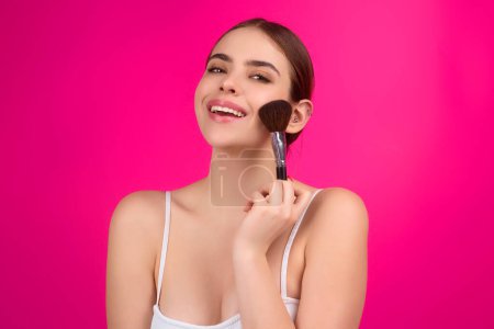 Téléchargez les photos : Young woman applying foundation powder or blush with makeup brush. Facial treatment, perfect skin, natural make up, facial beauty. Isolated on studio background. Applying makeup - en image libre de droit