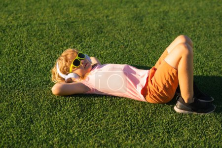 Téléchargez les photos : Les enfants aiment écouter de la musique dans les écouteurs, s'allonger sur l'herbe, rêver et se détendre. Enfant drôle émotionnel en t-shirt dans un casque dans un parc d'été. Concept de musique pour enfants - en image libre de droit