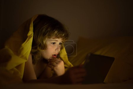 Photo for Child in bed under a blanket plays on a tablet in a game in the dark. The childs face is illuminated by a tablet. Child using tablet in bed at home. Kid in bedroom watching movie or or playing game - Royalty Free Image