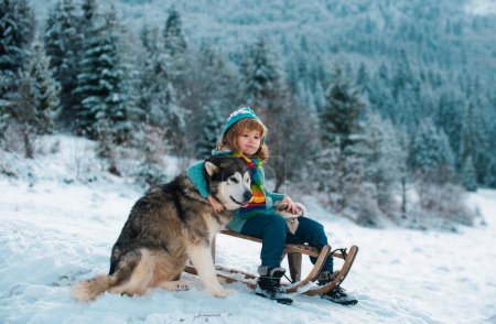 Foto de Boy kid enjoying a sleigh ride. Child on sleigh. Child plays outside in the snow. Winter, holiday and Christmas time. Kids hug embrace dog husky - Imagen libre de derechos