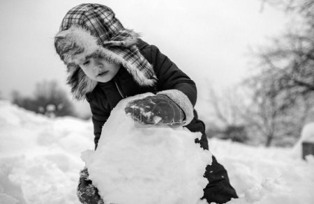 Téléchargez les photos : Beauté Enfant d'hiver avec bonhomme de neige dans le parc hivernal givré. Enfants d'hiver dans le parc hivernal givré. Portrait de petit garçon enfant dans la neige Jardin faire grosse boule de neige - en image libre de droit