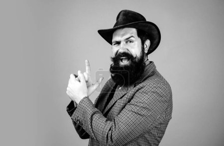 Photo for Young handsome bearded carefree funny man in suit with fingers like guns. Close up photo of fine brunet in hat with imagine gun in hands on isolated background - Royalty Free Image