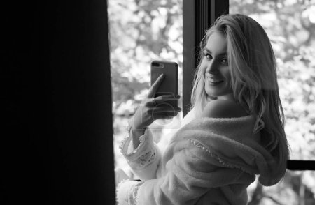 Photo for Morning of beautiful young woman with phone near window. Chilling woman in pajama laughing at home - Royalty Free Image
