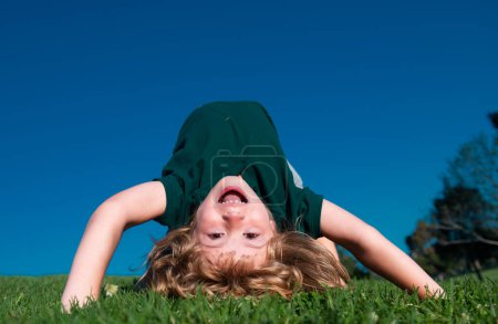 Téléchargez les photos : Joyeux enfant jouant à l'envers sur l'herbe verte. Enfant drôle en plein air dans le jardin de printemps. Journée des enfants. Joyeux concept d'enfance insouciante - en image libre de droit