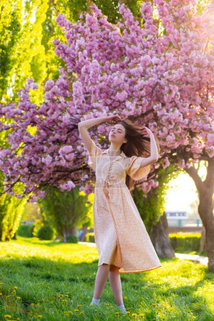 Foto de Una joven cerca del parque Blossom. Chica de primavera. Mujer en el árbol floreciente de primavera. Hermosa mujer joven en traje de estilo verano con flores al aire libre. Parque de sol de primavera. Belleza mujer disfrutando del olor en un - Imagen libre de derechos