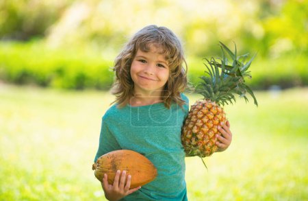 Téléchargez les photos : Enfant garçon tenant ananas et noix de coco souriant avec un visage heureux. Fruits d'été - en image libre de droit