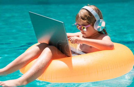 Téléchargez les photos : Voyage d'été d'affaires. Joyeux petit garçon apprenant ordinateur portable en ligne. Enfant se détendre sur anneau flottant en utilisant un ordinateur dans la piscine. Affaires d'été et technologie - en image libre de droit