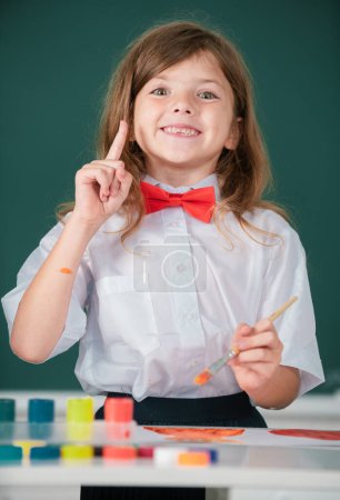 Téléchargez les photos : Child girl draws in classroom sitting at a table, having fun on school blackboard background. Portrait of little girl smiling happily while enjoying art and craft drawing lesson - en image libre de droit