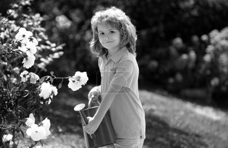 Foto de Lindo niño rizado niño en un vestido de verano que trabaja en el jardín regando flores. Jardinería para niños. Niños trabajando en el patio trasero. Niño con agua puede - Imagen libre de derechos