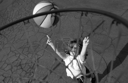 Téléchargez les photos : Petit garçon mignon en uniforme de basket-ball sautant avec une balle de basket pour tirer. Enfant heureux jouant au basket sur le terrain de jeu - en image libre de droit