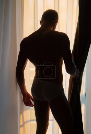 Téléchargez les photos : Un beau jeune homme torse nu qui regarde par la fenêtre. Sexy gay silhouette sur fenêtre ombre - en image libre de droit