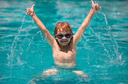 Téléchargez les photos : Enfant garçon avec les pouces levés mains levées nager dans la piscine. éclaboussures dans l'eau en s'amusant loisirs à bras ouverts - en image libre de droit