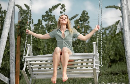 Photo for Outdoor portrait of a smiling happy girl. Fashion woman on wooden bench. Beauty blonde girl relaxing outdoors on porch swing in the nature. Summer mood. Happy cheerful girl laughing at park - Royalty Free Image