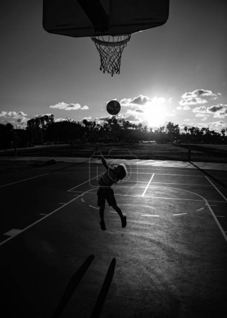Téléchargez les photos : Petit garçon mignon sautant avec une balle de basket pour tirer. Petit garçon jouant au basket. Silhouette de style de vie actif pour enfants au coucher du soleil - en image libre de droit