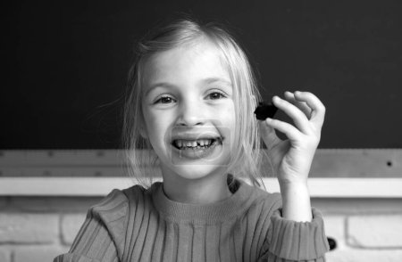 Téléchargez les photos : Gros plan portrait de souriante petite fille heureuse assise sur le bureau de la table dans la salle de classe, mangeant du chocolat. Petite drôle de visage d'écolière - en image libre de droit