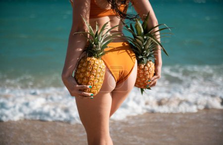Photo for Female closeup buttocks in thongs bikini, sexy ass. Young woman holding a pineapple on sea sand beach background - Royalty Free Image