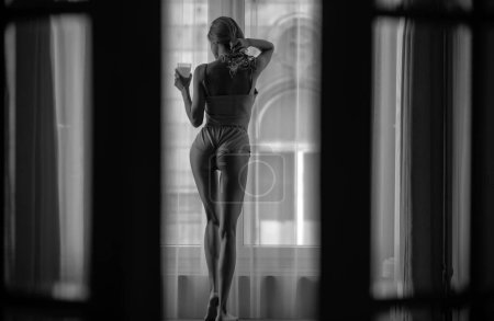 Téléchargez les photos : Belle femme sexy avec un corps sexy près de la fenêtre à la maison. Vue arrière des jeunes femmes en bonne santé en forme dans la chambre. Jus de boisson du matin - en image libre de droit