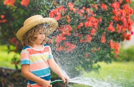 Téléchargez les photos : Un garçon heureux verse de l'eau dans un tuyau. Enfant arrosant des fleurs dans le jardin. Jardinage à domicile - en image libre de droit