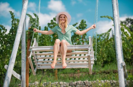 Foto de Retrato al aire libre de una chica feliz sonriente. Hermosa joven mujer se balancea en un columpio y disfrutar del hermoso clima de verano en el jardín natural. Mujer relajada y descansando en vacaciones de verano - Imagen libre de derechos