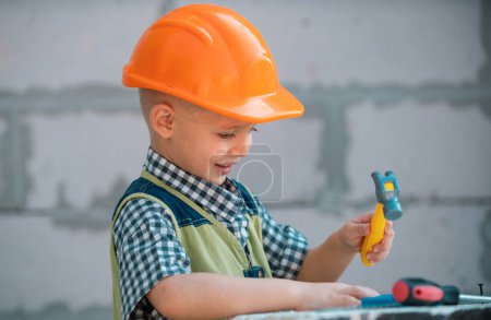 Téléchargez les photos : Enfant en casque dur tenant le marteau. Petit enfant aidant avec des outils de jouet sur le site construciton. Des enfants avec des outils de construction. Travailleur de la construction. Réparation maison - en image libre de droit