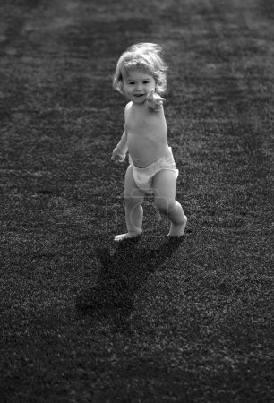 Foto de Lindo bebé divertido niño aprendiendo a gatear paso, divertirse jugando en el césped en el jardín en pantalones de pañales - Imagen libre de derechos