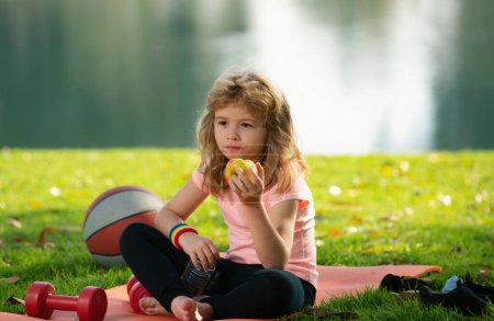 Téléchargez les photos : Fatigué adolescent garçon de sport manger de la pomme après l'entraînement dans le parc d'été. Concept de fitness et mode de vie sain. Concept de sport pour enfants. Protection des soins médicaux - en image libre de droit