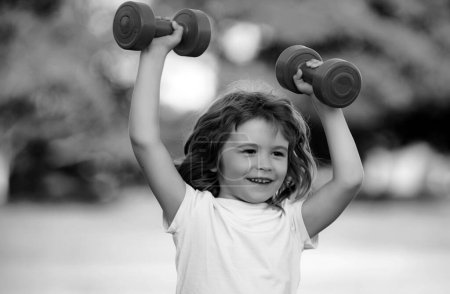 Foto de Feliz niño emocionado haciendo ejercicios con pesas en el parque verde. Retrato de primer plano de niño sonriente deportivo con mancuernas. Niño feliz ejercicio al aire libre - Imagen libre de derechos