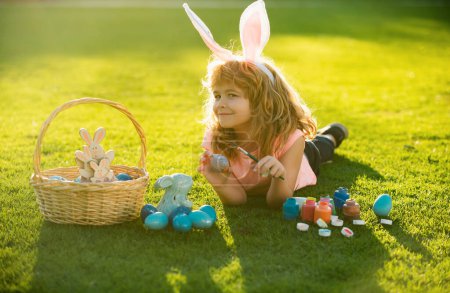 Foto de Niño con traje de conejo con orejas de conejo cazando huevos de Pascua en la hierba en el parque de primavera. Los niños de Pascua con orejas de conejo y sobre el fondo de hierba - Imagen libre de derechos