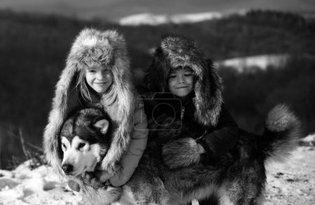 Téléchargez les photos : Petits enfants drôles embrassé chien husky en hiver en plein air. Souriant enfants amis étreindre chien dans le gel journée enneigée en plein air. Deux beaux garçons une fille se reposant ensemble dans le parc avec fond de neige - en image libre de droit