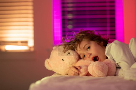 Photo for Cute drowsy sleepy child with toy teddy bear in bed, bedtime, childhood and growth kids concept, close-up indoor portrait. Child is afraid of the dark. Nightmares and terrible dreams in children - Royalty Free Image