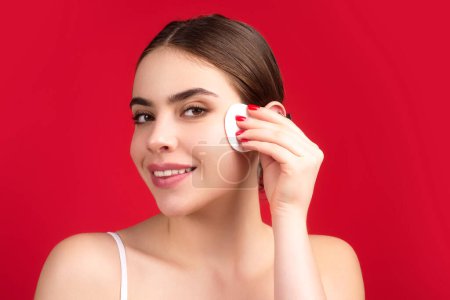 Téléchargez les photos : Beautiful young woman with cotton pad removing makeup. Skin care. Studio portrait of girl cleaning her face with cotton pad, isolated beige background. Face toner and cotton pad - en image libre de droit