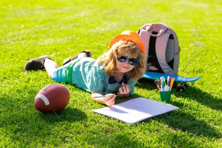 Photo for Child with artwork homework on playground. Clever school boy doing homework, writing on copy book in green grass of park. Back to school, knowledge, education, learning concepts - Royalty Free Image