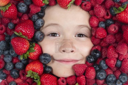 Téléchargez les photos : Une saine alimentation. Les enfants font face avec des fruits et des baies. Fraise d'été, myrtille, framboise, fond de mûre. Vue du dessus photo du visage de l'enfant en arrière-plan de baies. Enfants en bonne santé manger - en image libre de droit