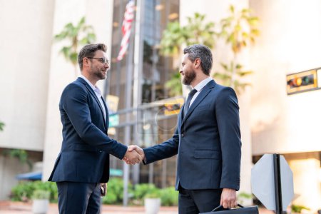 Photo for American businessman shaking hands with partner. Handshake between two business men. Two businessmen shaking hands on city street. Business men in suit shaking hands outdoors. Business team - Royalty Free Image
