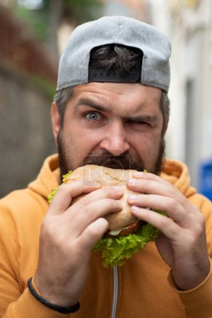 Foto de Hambre Hombre comer hamburguesa al aire libre. El hombre come sabrosa hamburguesa en la calle. Hamburguesa en el almuerzo. Hamburguesa con queso o hamburguesa. Hombre comiendo sabrosas hamburguesas afuera. Sabrosa hamburguesa. Barbudo hombre disfrutando de deliciosa hamburguesa - Imagen libre de derechos