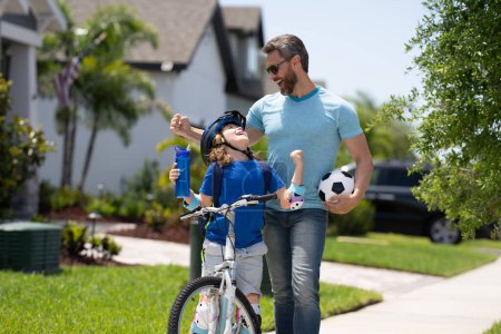 Photo for Father teaching son riding bike. Dad helping child son to ride a bicycle in american neighborhood. Child in bike helmet learning to ride cycle with father. Happy fathers day. Summer sport with child - Royalty Free Image