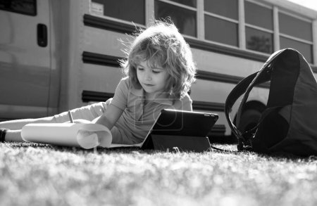Foto de De vuelta a la escuela. Feliz estudio infantil en el parque cerca del autobús escolar. Colegial haciendo tarea o educación en línea al aire libre - Imagen libre de derechos
