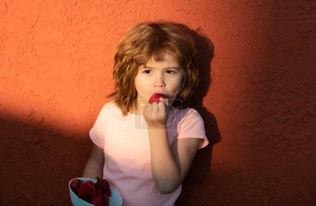 Téléchargez les photos : Drôle de visage de mignon petit garçon mangeant une fraise - en image libre de droit